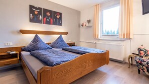 Camera da letto con un letto in legno, due cuscini, un comodino e un tavolino, una finestra con tende e decorazioni alle pareti.