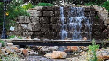 Ein Wasserfall stürzt über eine Steinmauer in einen Teich, umgeben von Steinen und Grünpflanzen.
