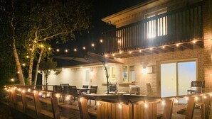 Una terraza de madera con luces colgantes, mobiliario exterior y un edificio de ladrillo con grandes ventanales.