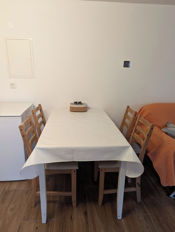A compact room with a white table, four wooden chairs, a small round table with two cups, a bed with an orange blanket, and a white wall with a thermostat.