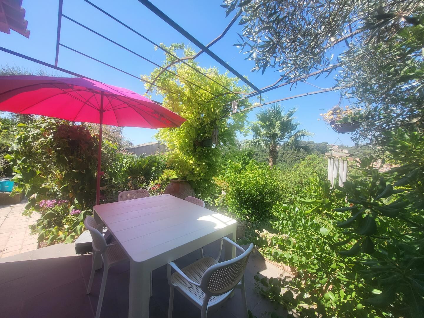Eine Terrasse mit weißem Tisch und Stühlen, einem roten Sonnenschirm und üppigem Grün.
