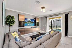 A modern living room with a grey sofa set, a flat-screen TV mounted above a wooden coffee table, and a large window with a view of a pool and greenery. - Spacious 6-bedroom house in beautiful Kissimmee with AC (Kissimmee)