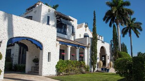 Un edificio de estuco blanco con detalles azules, un porche cubierto y un jardín bien cuidado con palmeras.