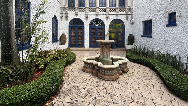 Un patio con fuente, edificios blancos con puertas azules y un sendero adoquinado.