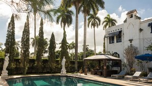 Una piscina con sillas de descanso y un pabellón, rodeada de palmeras y un edificio blanco con detalles azules.