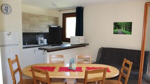 A dining area with a round wooden table, four wooden chairs, a small decorative item, a clock on the wall, a kitchen area with cabinets and appliances, and a framed picture on the wall. - Les Faons - ROUBION - Gîte (ROUBION)