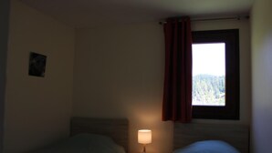 A bedroom with two beds, a nightstand with a lamp, a window with a view of trees, and a curtain. - Les Faons - ROUBION - Gîte (ROUBION)