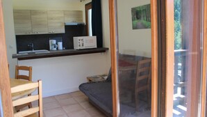 A small kitchenette with a sink, coffee maker, and microwave. There is a wooden chair and a small table with a magazine. A sliding glass door leads to an outdoor area with a view of a forest. - Les Faons - ROUBION - Gîte (ROUBION)