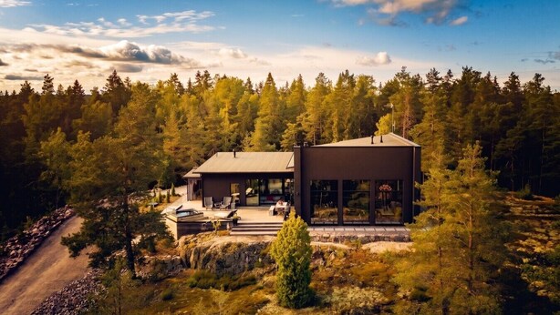 Une cabane moderne à deux étages, dotée d’une grande façade vitrée et entourée d’un paysage boisé.