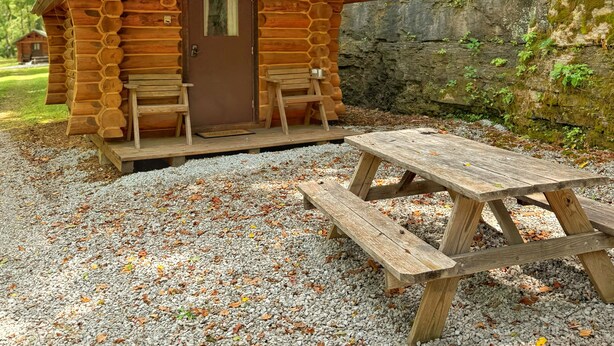 Une cabane rustique avec une façade en bois, une porte d’entrée, deux bancs et une table de pique-nique devant.