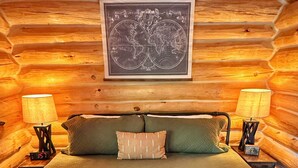 Une chambre à coucher aménagée dans un chalet en rondins, avec un lit, deux lampes de chevet, une carte du monde accrochée au mur et un tapis aux motifs rouges et blancs.