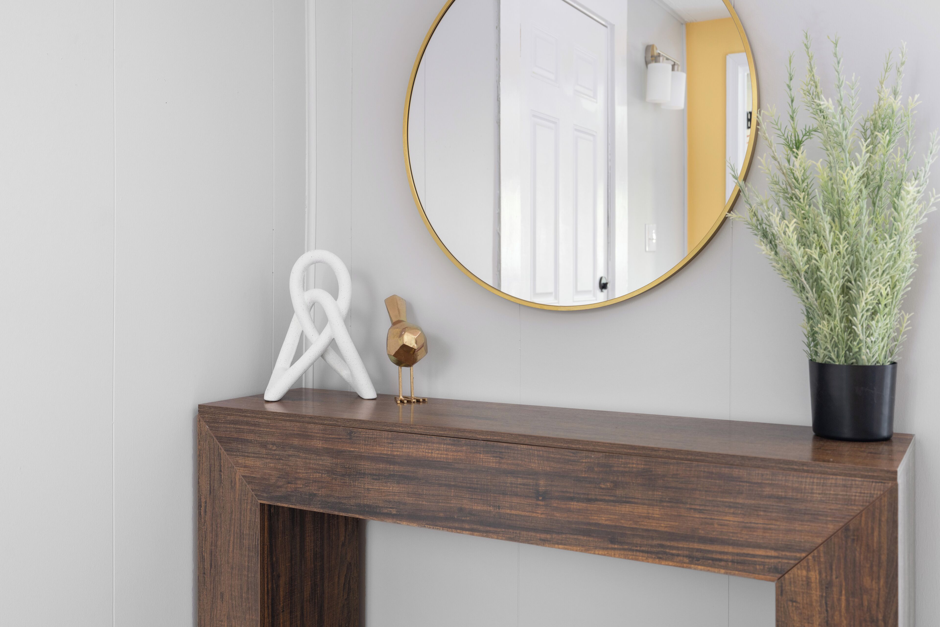 Une console en bois, surmontée d’un miroir rond, avec une plante en pot et un objet blanc en forme de ruban posé sur le plateau.