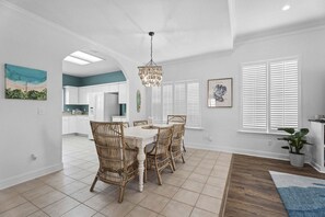 Un coin repas avec une table blanche et des chaises en osier, un lustre et une vue sur la cuisine à travers une arche.