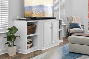 Una sala de estar con un televisor de pantalla plana sobre un mueble blanco, una planta en maceta, una cesta de mimbre y un sofá con una manta.
