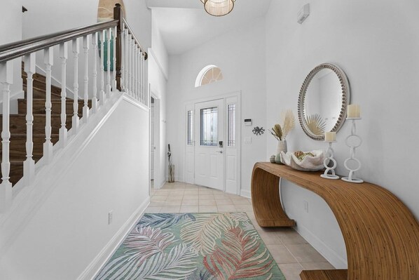 Un couloir avec un escalier en bois, une porte blanche, un miroir et une console ornée d’objets décoratifs.