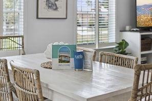 Una mesa de comedor con una caja de bienvenida, una lata y sillas de mimbre en una habitación con televisión y persianas.