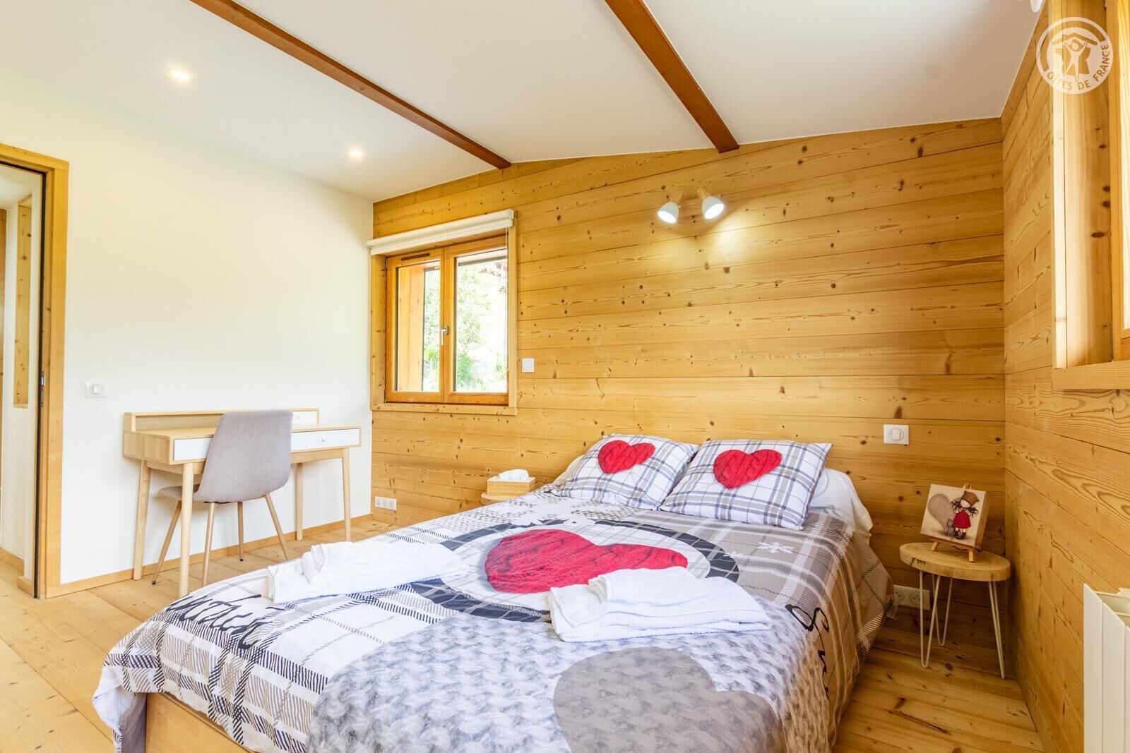 A cozy bedroom with a wooden ceiling, a bed with a patterned bedspread, a desk with a chair, and a window.