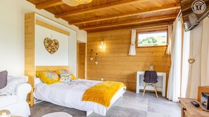 A cozy bedroom with a bed, a desk, and a chair. There is a window with curtains, a wooden ceiling, and a wall-mounted TV.