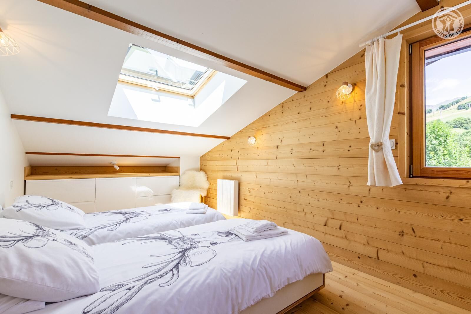 A bedroom with two beds, wooden walls, a skylight, and a view of greenery.