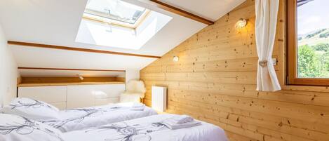 A bedroom with two beds, wooden walls, a skylight, and a view of greenery.