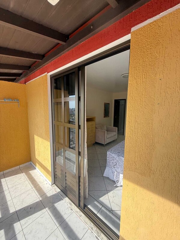 Una habitación con puerta de vidrio corredera que da a un balcón, una cama con cubrecama blanco y una mesita de noche pequeña.
