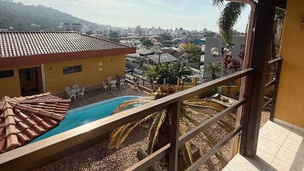 A balcony with a view of a swimming pool, a building with a tiled roof, and a distant cityscape.