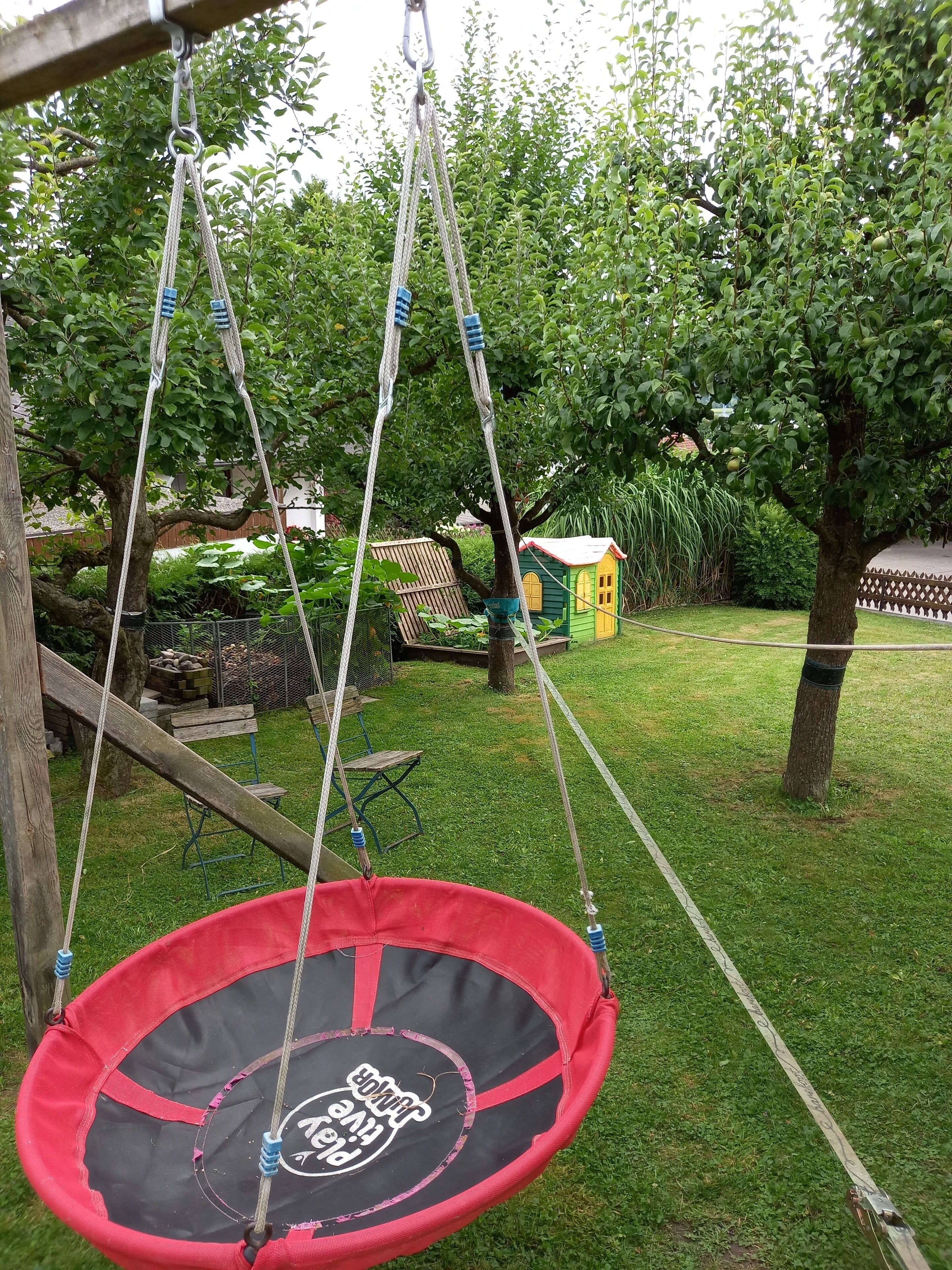 Un trampolín rojo con un logotipo, suspendido por cuerdas en un césped con árboles y una casa pequeña al fondo.