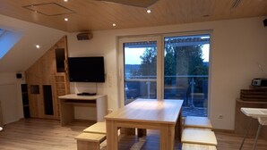 A modern, well-lit room with a wooden floor, a large table with benches, a TV, and a sliding glass door leading to an outdoor area.