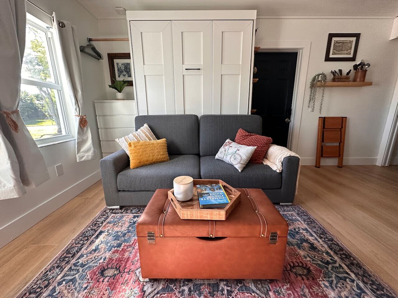 Ein Wohnzimmer mit grauem Sofa, einem braunen Ledersitzhocker, einem gemusterten Teppich und einem Fenster mit Vorhängen.