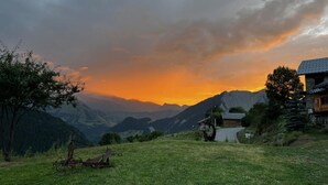 Un chalet de montagne rustique, avec une façade en pierre et une terrasse en bois, entouré d’une végétation luxuriante et offrant une vue sur les montagnes lointaines.