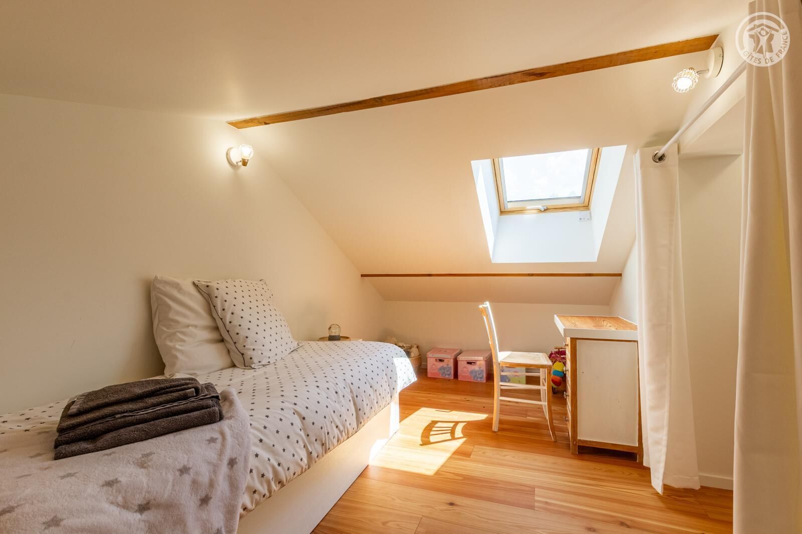 A bedroom with a sloped ceiling, a skylight, a bed with a star-patterned duvet, a desk with a chair, and a small table with toys.