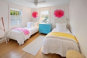 A bedroom with two beds, a window with blinds, a ceiling fan, and a decorative wall piece. - Modern Saltbox in Edgartown Village (Edgartown)