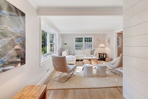 Un salon spacieux avec un canapé blanc, deux fauteuils blancs, une table basse et une grande fenêtre équipée de stores.