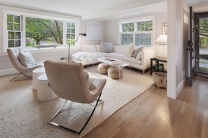 Ein modernes Wohnzimmer mit einem beigefarbenen Sofa, einem weißen Sessel und einem Couchtisch. Große Fenster bieten Blick auf Bäume, und ein Lampenschirm steht auf einem Beistelltisch.