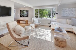 Ein modernes Wohnzimmer mit einem weißen Sofa, einem Schaukelstuhl, einem Couchtisch und einem großen Fenster mit Blick auf Grünflächen.