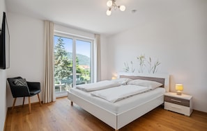A modern bedroom with a large bed, a chair, a nightstand, and a view of the outdoors.