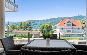 A balcony with wicker chairs and a table, overlooking a lake and a red-roofed building.