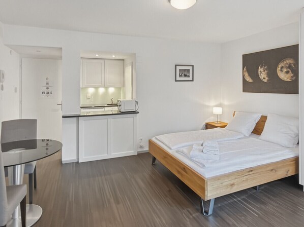 A compact hotel room with a wooden bed, a small kitchenette, a round dining table, and a wall-mounted picture of the moon phases.