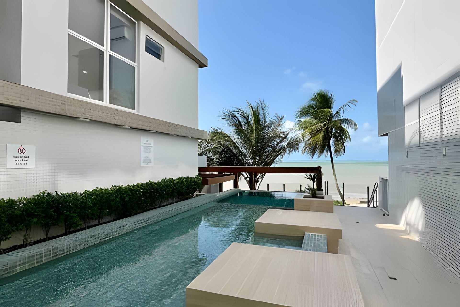 A modern pool area with clear water, surrounded by white walls and a view of the beach and palm trees.