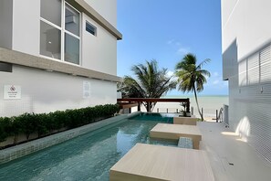 Un área de piscina moderna con agua cristalina, rodeada de muros blancos y vistas a la playa y palmeras.