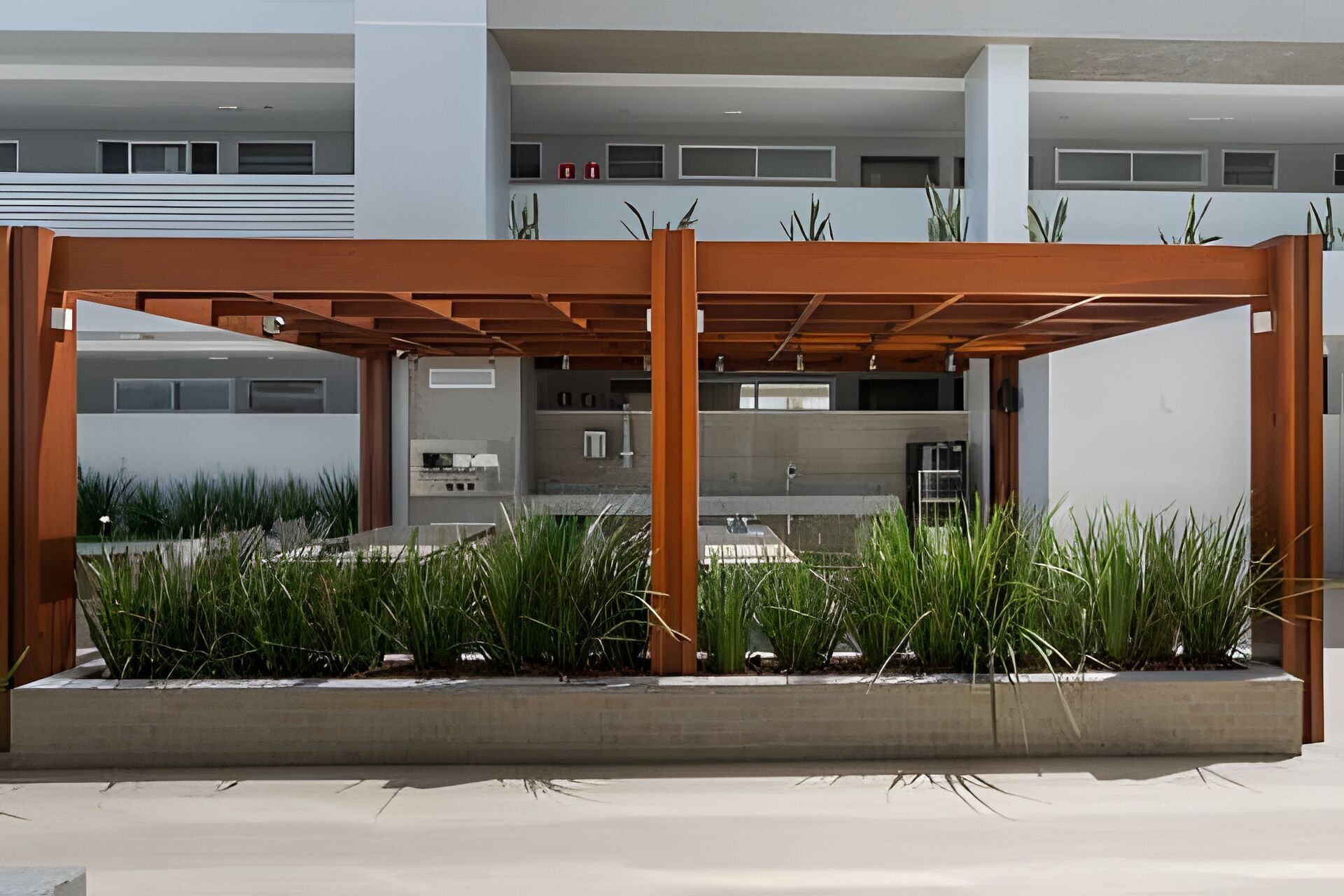 A modern outdoor seating area with a wooden pergola, surrounded by tall grasses, in front of a building with multiple windows and a visible kitchen area.