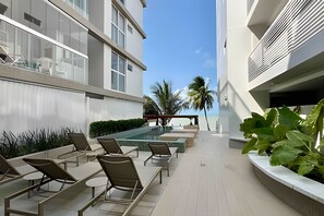 Un área lounge moderna al aire libre con tumbonas y piscina, rodeada de edificios y palmeras.