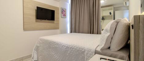 Quarto de hotel com cama, televisão fixada em painel de madeira, mesa de cabeceira com controle remoto e guarda-roupa.
