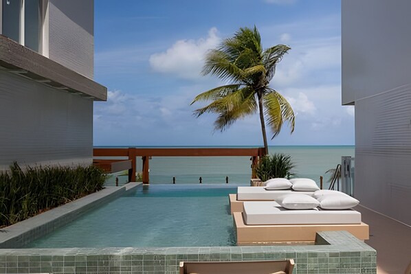 Zona de piscina con vista al océano, una palmera y sillas de descanso con cojines.