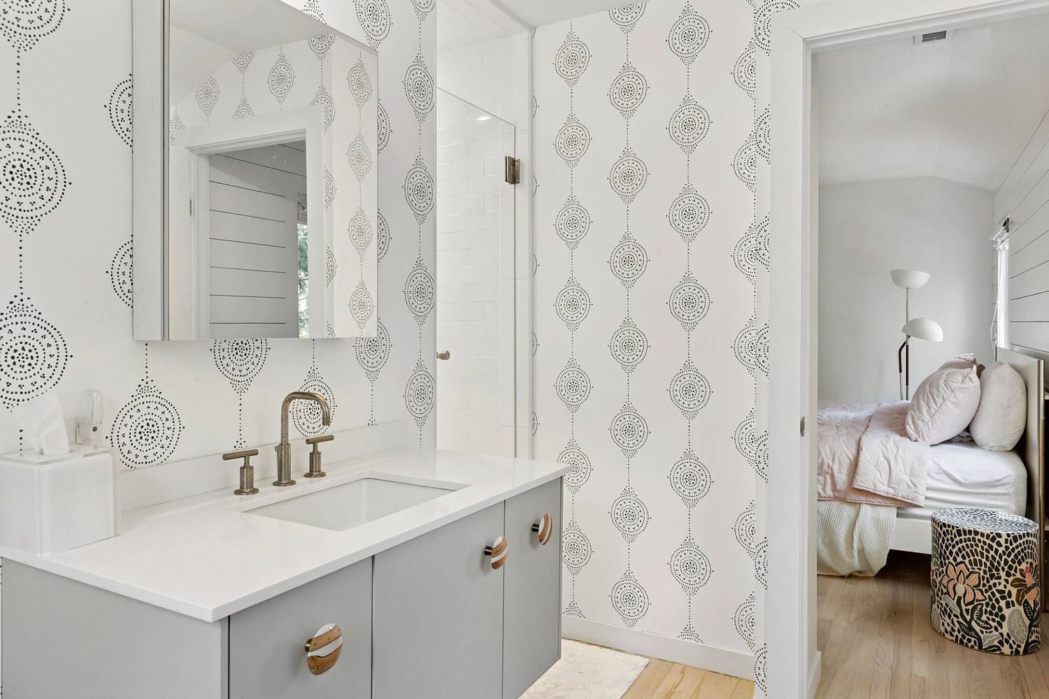A modern bathroom with a white sink, two faucets, and a mirror. The walls have a geometric pattern. There is a bed with white bedding and a lamp in the adjacent room.