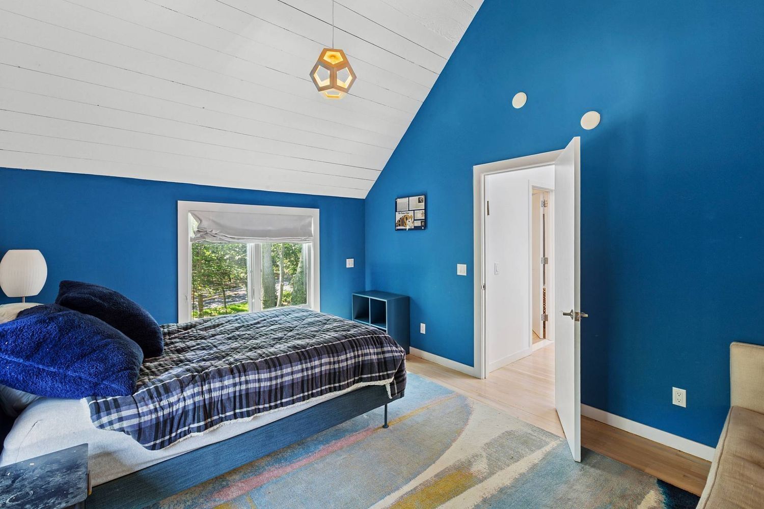 A bedroom with a large bed, a window with a view of greenery, a door, and a ceiling light.
