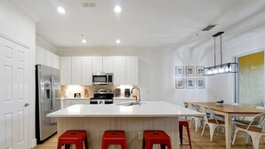Eine moderne Küche mit weißen Schränken, einem Edelstahl-Kühlschrank, einer Mikrowelle und einem Spülbecken. Vor der Kücheninsel stehen drei rote Hocker.