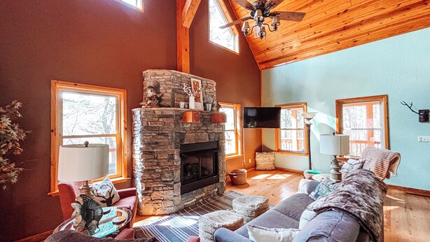 A cozy living room with a stone fireplace, wooden ceiling beams, and a large window.