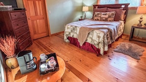 A cozy bedroom with a wooden ceiling, a bed with a plaid bedspread, a dresser, a coffee maker, and a small table with a cup and saucer.