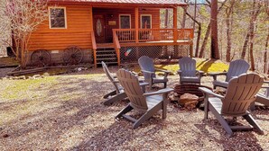 A wooden cabin with a red roof, a porch with Adirondack chairs, and a gravel ground.